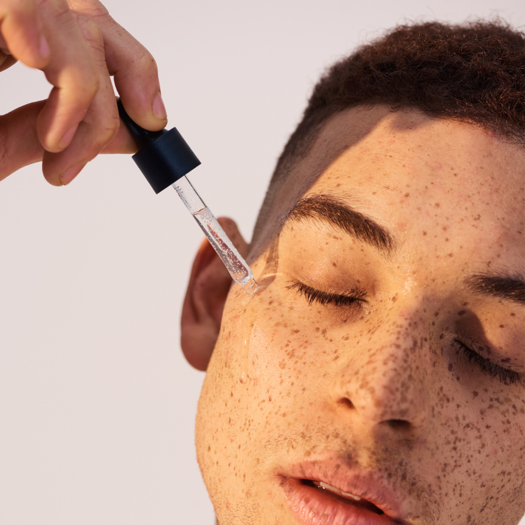 Man with freckles applying serum to left cheek with eyes closed